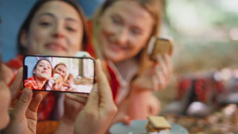 woman taking video of friends on camping holiday in forest lying in tent eating s'mores