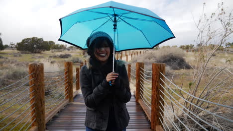 una hermosa joven sonriendo y riendo bajo la lluvia con un paraguas azul en la tormenta y el mal tiempo a cámara lenta