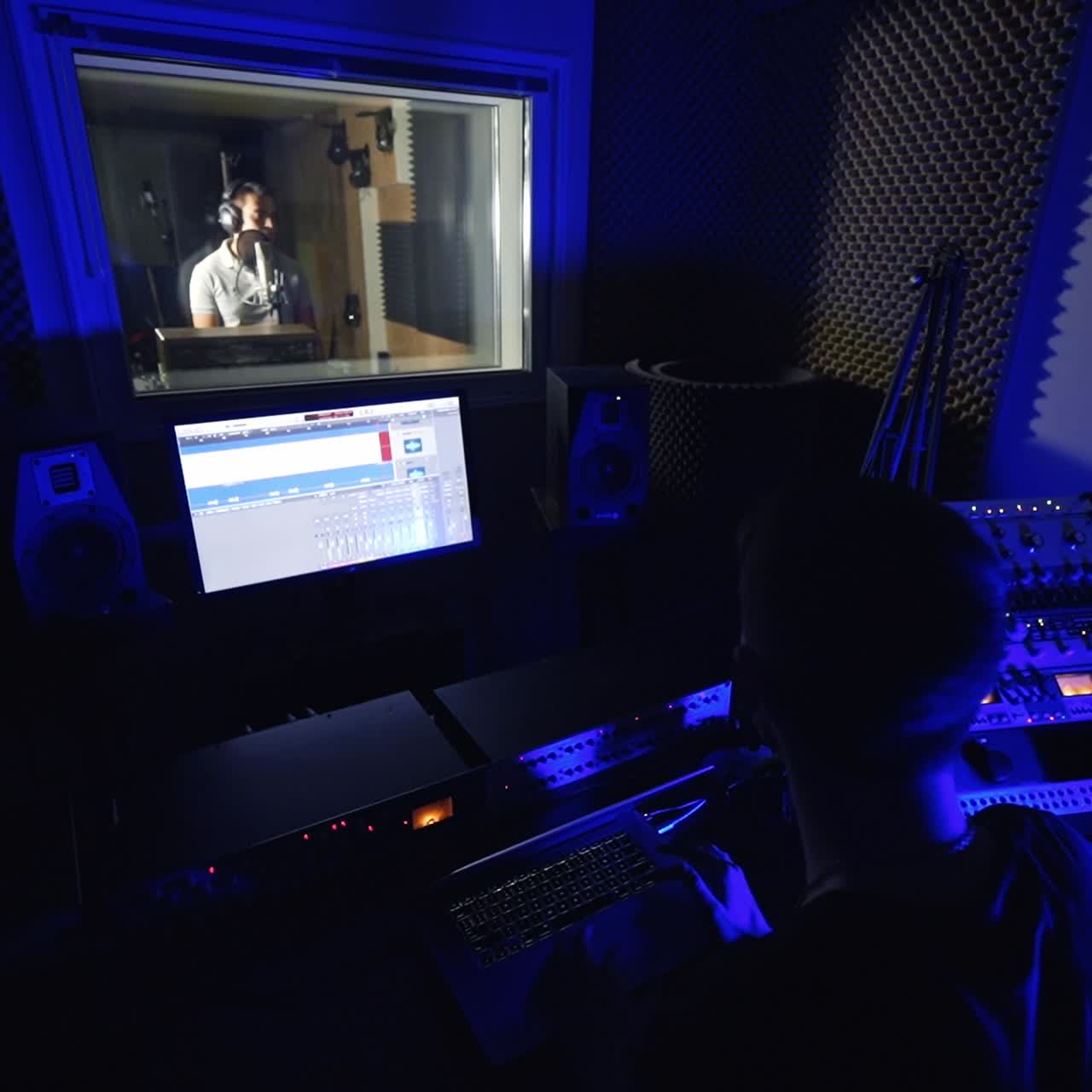 Music producer working in the little dark recording room. Sound engineer looks at computer screen. Male singer performing a song at backdrop