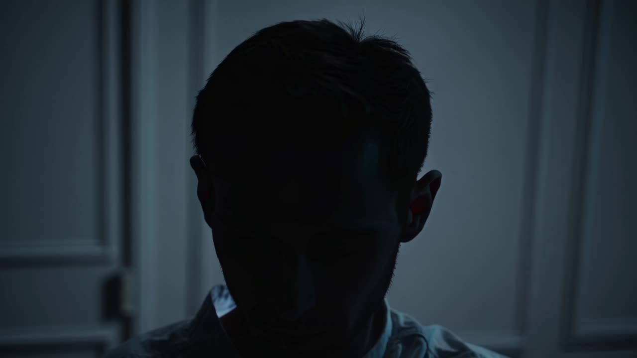 Young man is engrossed in his smartphone in a dimly lit room, his face illuminated by the bright glow of the screen, creating a dramatic contrast of light and shadow