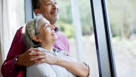 Feliz-Pareja-De-Ancianos-Birraciales-Abrazándose-En-La-Ventana-De-Casa