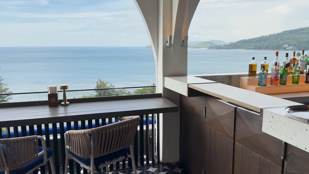 A serene clifftop bar in Phuket offers stunning ocean views, with a well-stocked counter and inviting seating under natural daylight