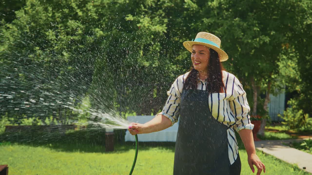 Woman watering garden with hose in sunny backyard, showcasing vibrant greenery and flowers