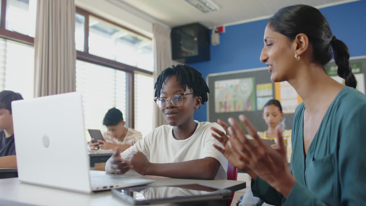 In school, teacher helping student with laptop in classroom, others studying