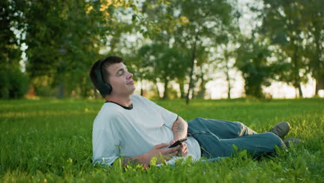 man in white shirt wearing headphones lying sideways on grassy field holding smartphone, nodding to music, surrounded by lush greenery and trees with sunlight streaming through background