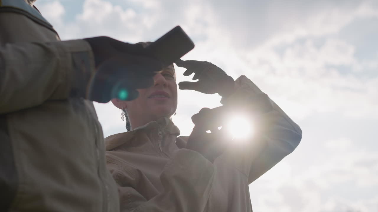 close up of woman shielding eyes from sun flare while holding mobile device, colleague gloved hand and phone visible in foreground, soft backlight creating lens flare