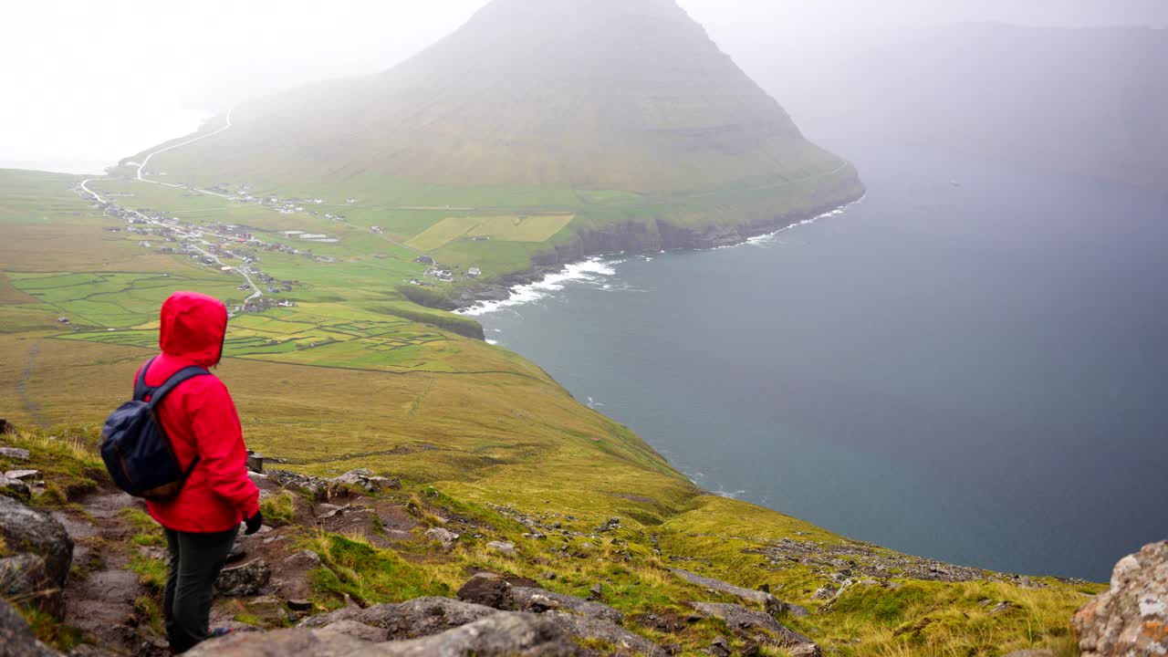 vista trasera de una persona con chaqueta de lluvia roja levanta los brazos mirando hacia abajo en el pueblo de vidareidi y el fiordo de las islas feroe