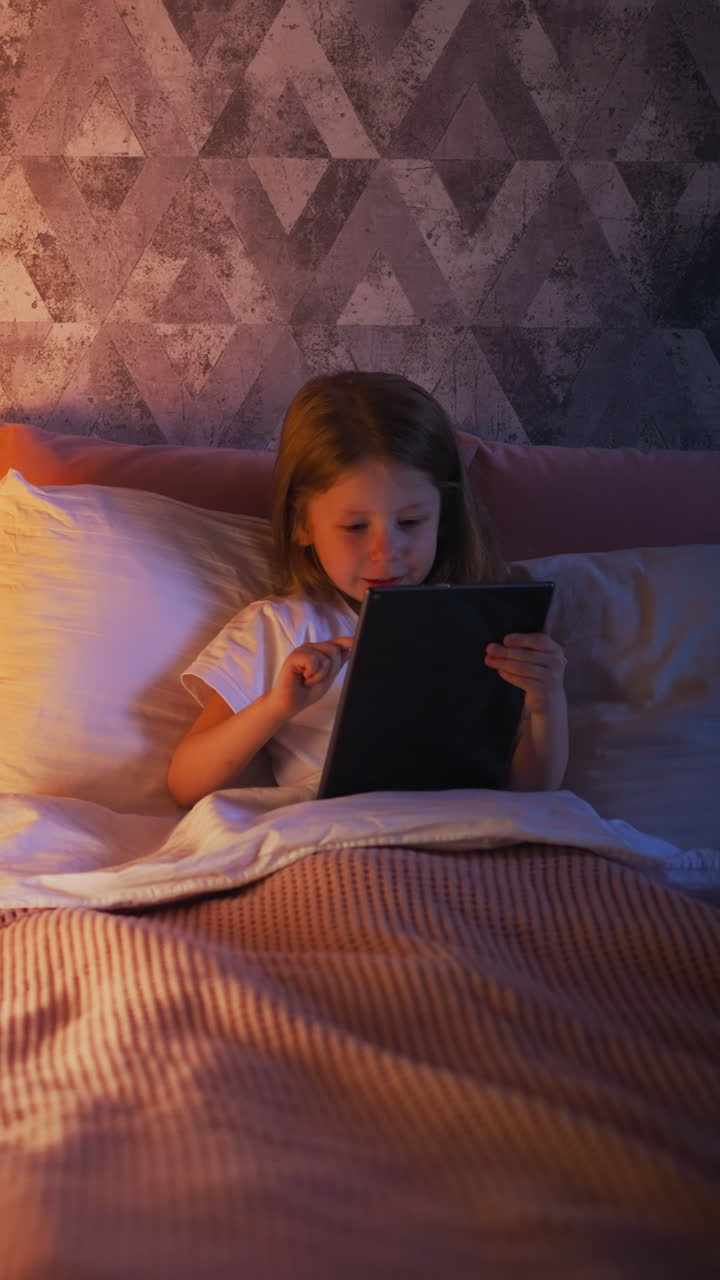 Smiling little kid surfs internet on tablet computer searching film in comfortable bed with soft pillows and covering near toy wigwam with glowing lamp slow motion
