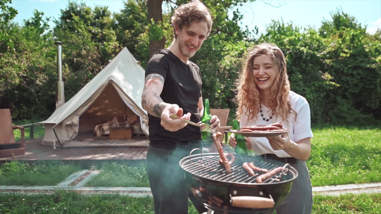 Young couple frying meat on the grill with beer, putting it on the plate and having fun. Greenery around. Glamping. Slow motion