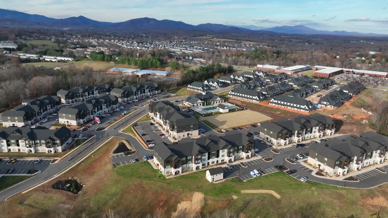 Modern luxury apartment community in America, with condominium buildings, parking areas, landscaped grounds, and community swimming pool with mountains in background. Aerial approaching shot. VA, U.S