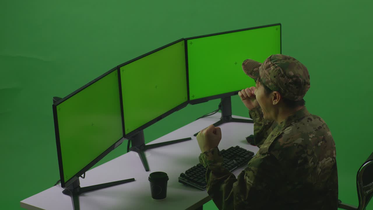 vista laterale di un felice soldato asiatico che celebra e balla mentre lavora con un simulacro di più monitor di computer in uno studio a schermo verde