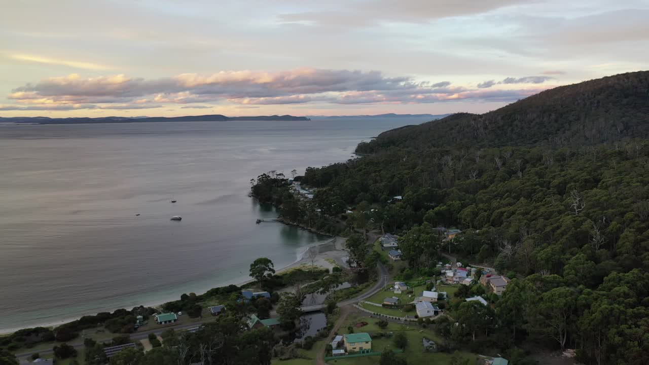 drone aéreo lento pan alrededor de la bahía de la isla durante la puesta de sol sobre la playa al atardecer, cielo púrpura y naranja y cordillera con pequeñas chozas y bosque con nubes