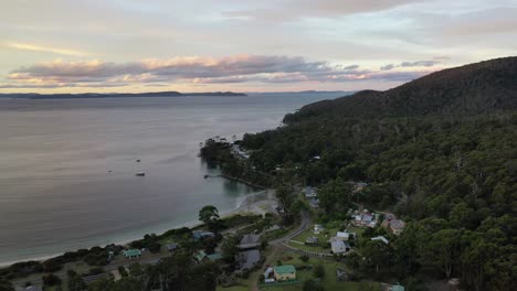 Slow-aerial-drone-pan-around-island-bay-during-early-sunset-over-beach-at-dusk,-purple-and-orange-sky-and-mountain-range-with-small-shacks-and-forest-with-clouds