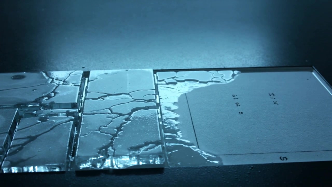 左から滴り落ちる透明な液体が、実験台のガラスタイルにあるひび割れに沿って広がり、滑らかな膜を形成している。