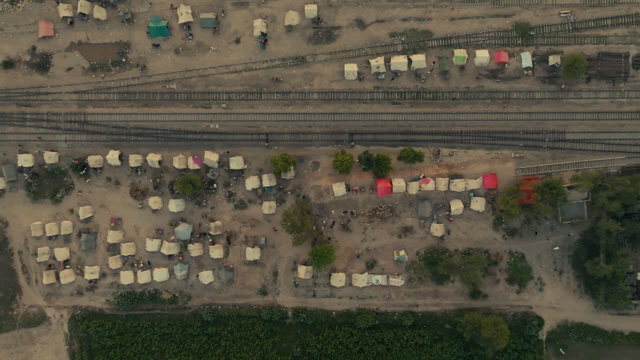 Bird's eye drone clip over large number of emergency tents and shelters set up directly on the dirt embankment flanking train tracks in a flood-affected agricultural area of Punjab, Pakistan