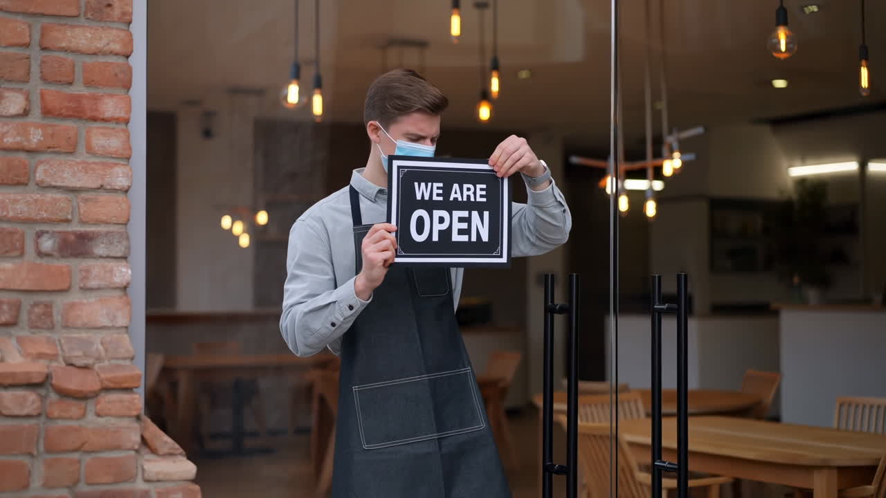 Young Man in Mask Puts 'We Are Open' Sign on Cafe Door