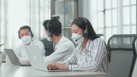 a woman of three asian call center agents wearing headset and mask speaking to customers on the call while two of her colleagues are talking during working at the office