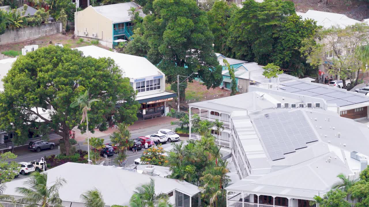 Drone footage captures a lush, tree-lined street with buildings in Port Douglas, Australia. Bright daylight enhances the vibrant greenery