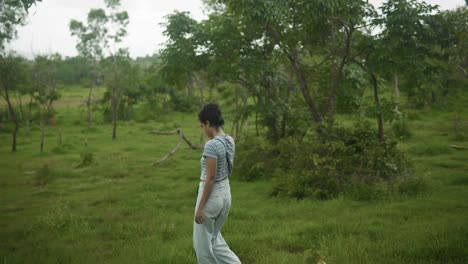 a girl, wearing a blue stripe shirt, pants, and carrying a black bag, walks through the grassy landscape with trees surrounding her