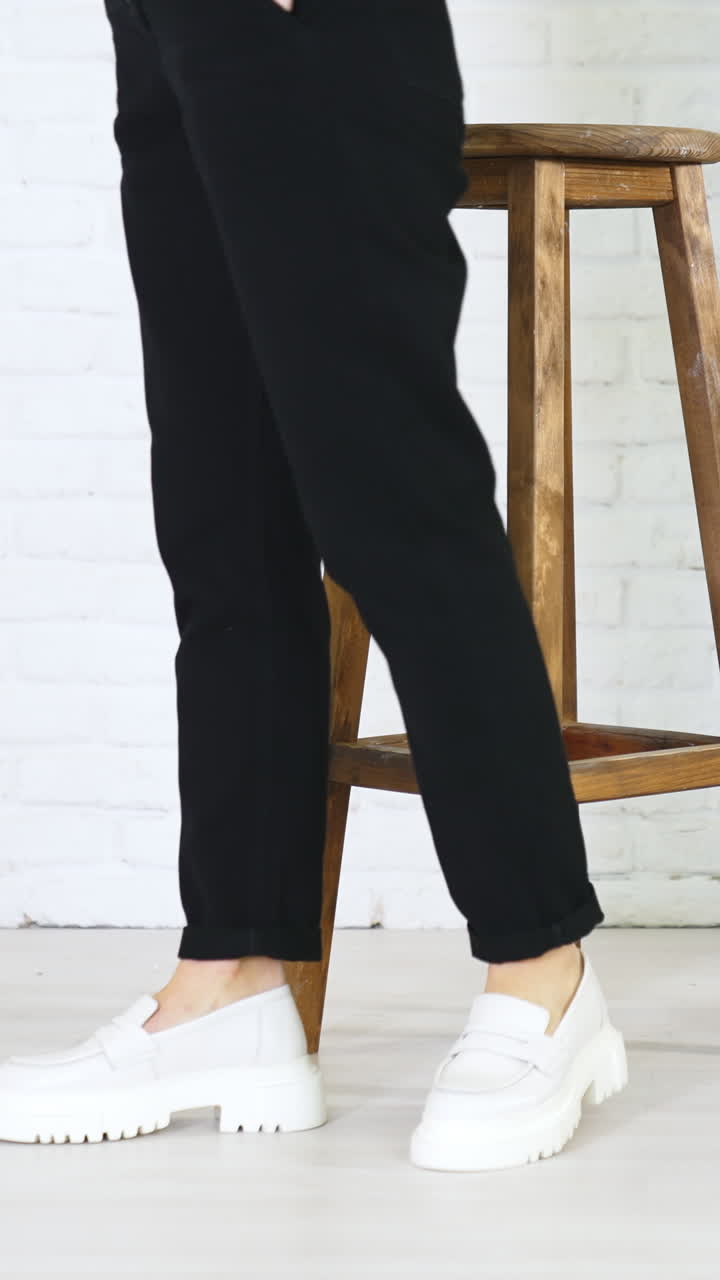 Female model posing in massive modern white shoes. Lady in black jeans walks in new pair of footwear in front of wooden stool. White wall backdrop. Vertical video