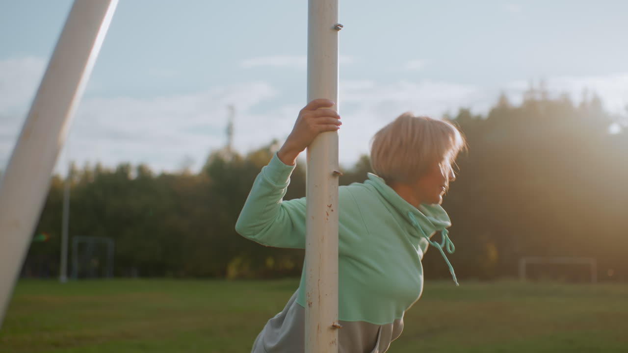 Sports woman holding poll while performing leg stretches front and back on green grassy field during sunny outdoor session surrounded by natural park scenery demonstrating fitness, and balance