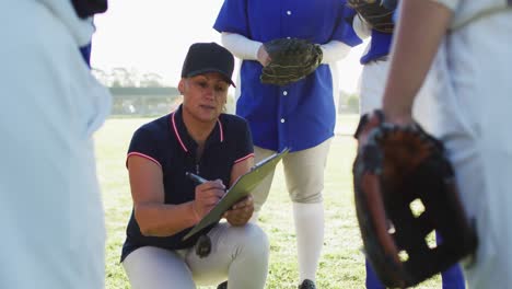 caucasian female baseball coach squatting writing on clipboard and talking to team on pitch