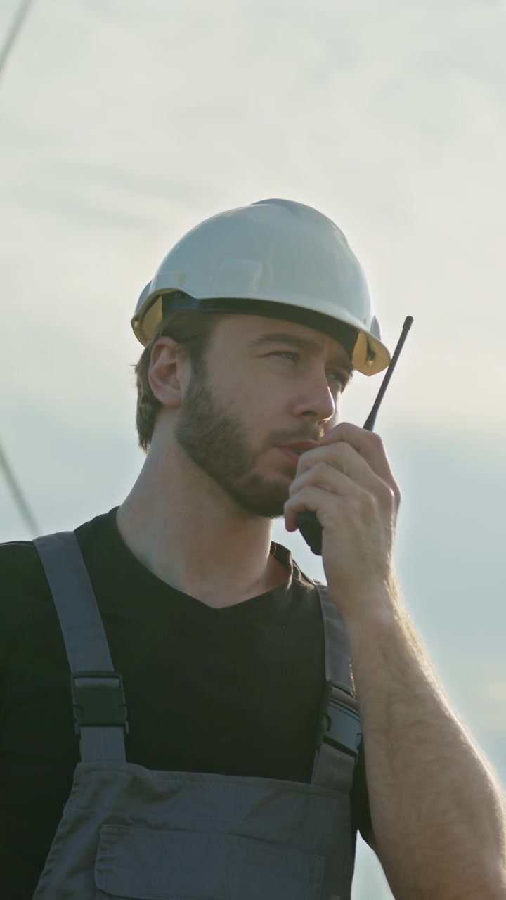 Uomo con cappello e tuta da lavoro usa una radio portatile per comunicare in un cantiere con turbine eoliche.