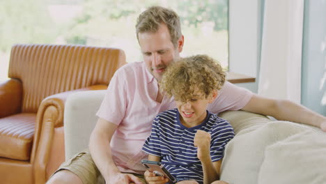 Padre-E-Hijo-Sentados-En-El-Sofá-En-Casa-Jugando-Juntos-Videojuegos-En-El-Teléfono-Móvil