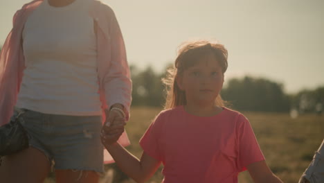 close up of girl child walking closely between two adults, holding their hands while moving through vast grassy field on sunny day, background features slightly blurred trees