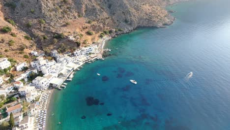 Drone-view-in-Greece-flying-over-blue-sea-in-Loutro-small-white-house-town-and-small-boats-next-to-a-hill-on-a-sunny-day