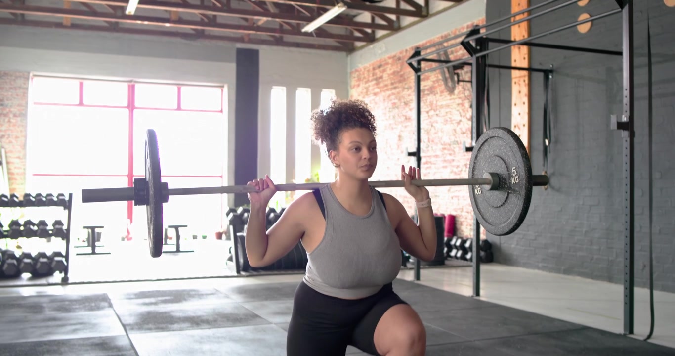 Donna afroamericana che regola la presa, attiva il core ed esegue affondi con bilanciere in palestra.