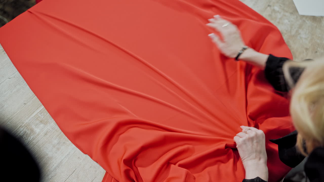 Femile tailor is layng out bright red fabric on the wooden table. Large piece of red material in the hands of a seamstress in workshop.