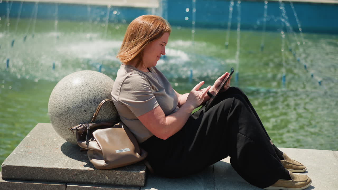 Loving woman sitting on stone bench near fountain operating phone with warmth and focus, relaxed mood under sunlight, gentle breeze over water, peaceful outdoor moment of quiet