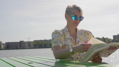 stylish young blonde woman reads a book, sitting at a picnic table, river in the background