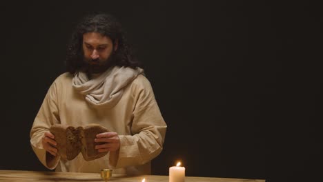 studio shot of man wearing robes with long hair and beard representing figure of jesus christ breaking bread