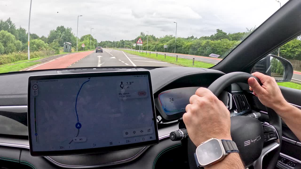 A person drives an electric vehicle along a rural road in Durham, England, interacting with a central touchscreen under natural daylight with steady camera perspective