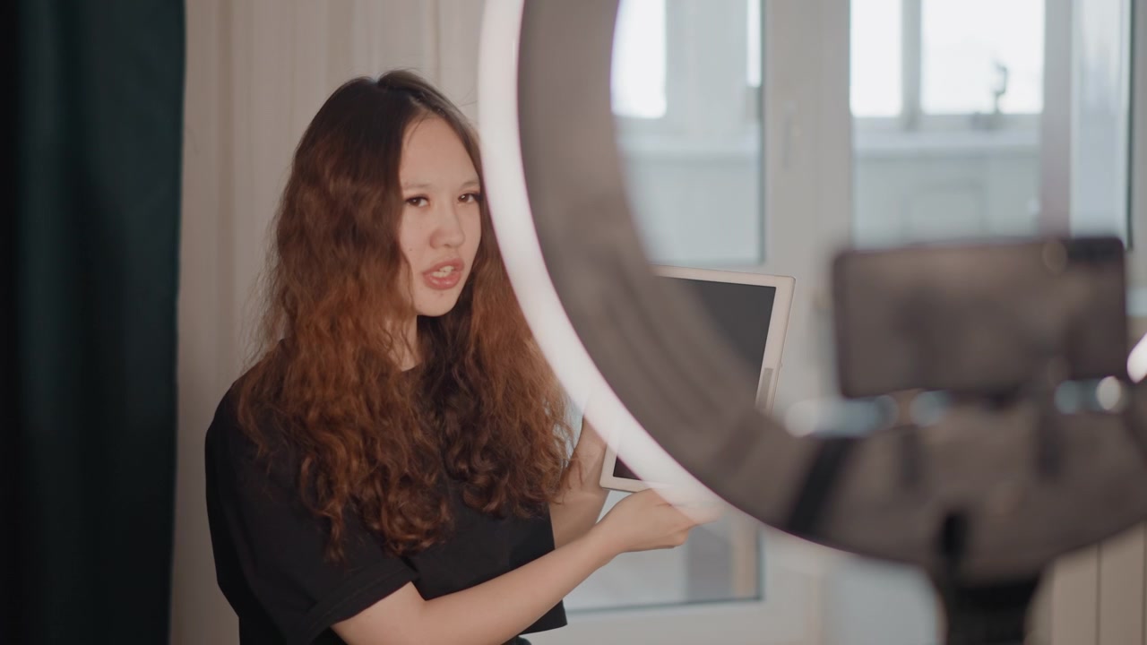 Mujer con cabello largo y rizado de pie en el interior cerca de una ventana luminosa, sosteniendo una tablet a la altura del pecho e inclinando la pantalla hacia la cámara en un claro gesto de presentación, con el rostro parcialmente cubierto por el cabello.