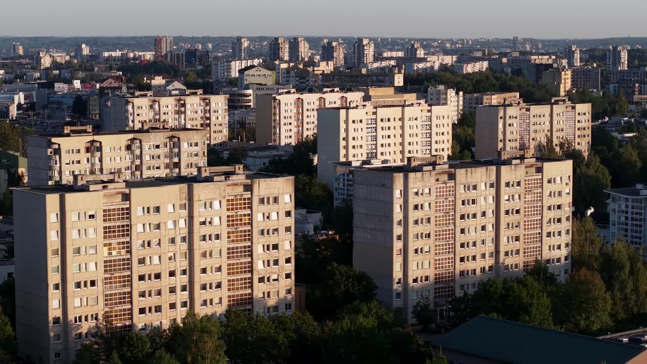 Дрон медленно пролетает над однотипными многоквартирными домами с отражениями заката, деревьями, городом и лесом на заднем плане