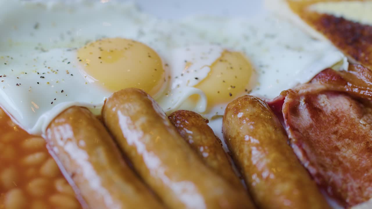 Close-up of fork and knife slicing sausage with eggs, bacon, and beans under natural lighting