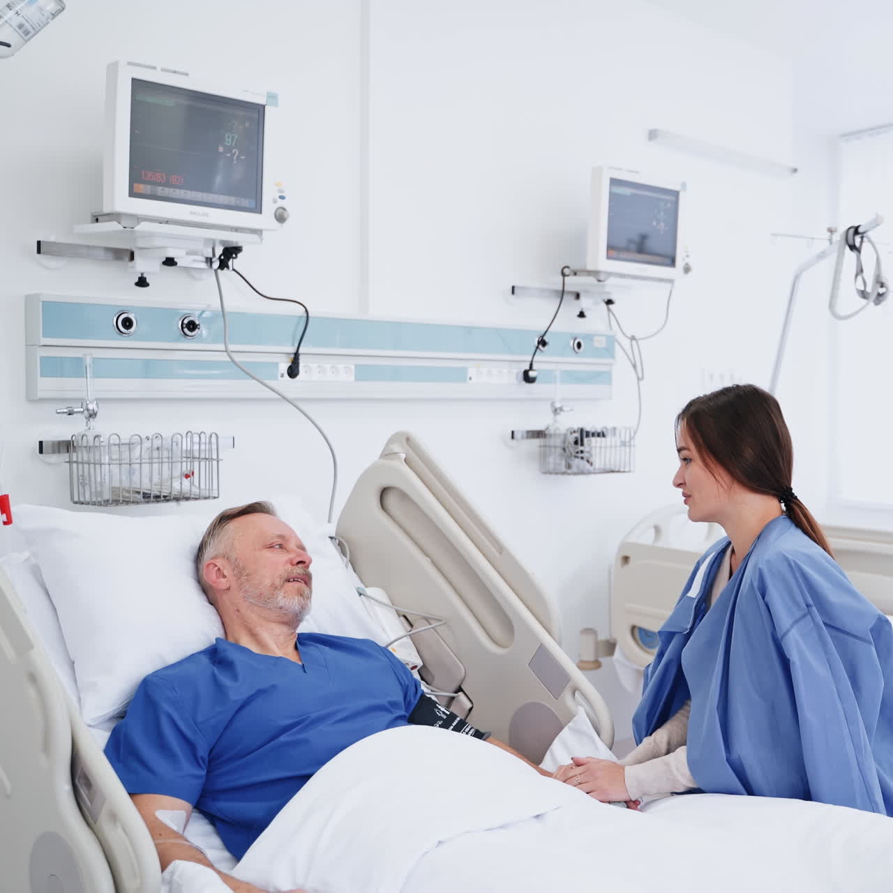 Visit to a patient in clinic. Young woman talks to her sick father. Senior man lying on a hospital bed and talking to his daughter in modern medical center.