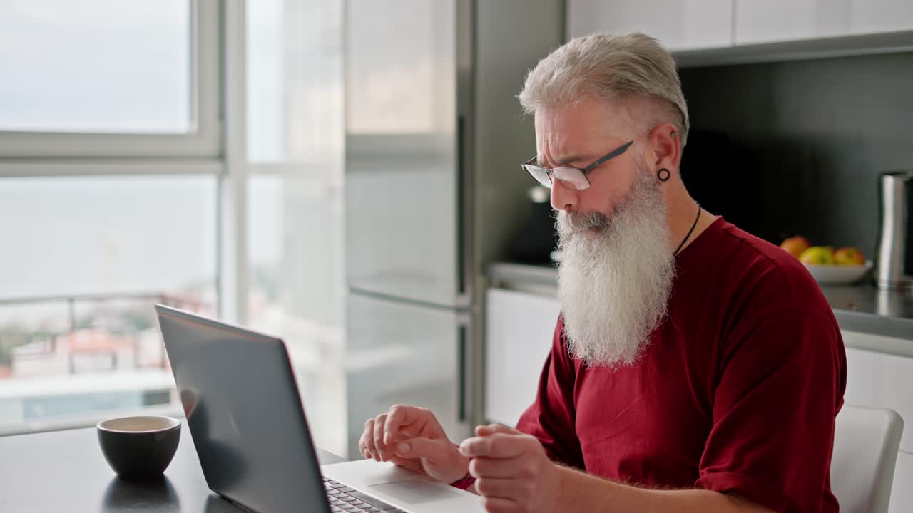 An elderly man with glasses with gray hair and a lush beard in a red shirt works on his gray laptop while sitting in the kitchen of a modern apartment
