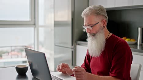 Ein-älterer-Mann-Mit-Brille,-Grauem-Haar-Und-üppigem-Bart-In-Einem-Roten-Hemd-Arbeitet-An-Seinem-Grauen-Laptop,-Während-Er-In-Der-Küche-Einer-Modernen-Wohnung-Sitzt