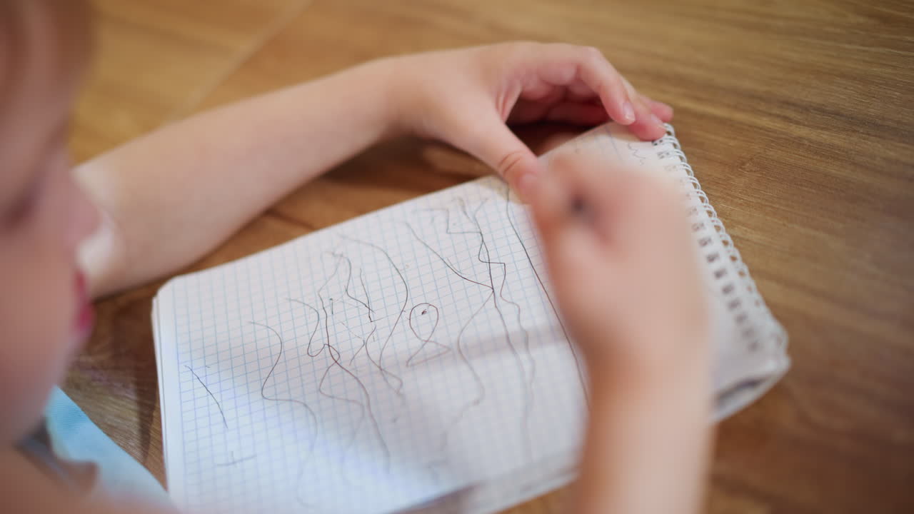 Child drawing random lines with pen on graph paper notebook at wooden table, focusing on creative activity, practicing hand coordination, exploring imagination, in casual home environment