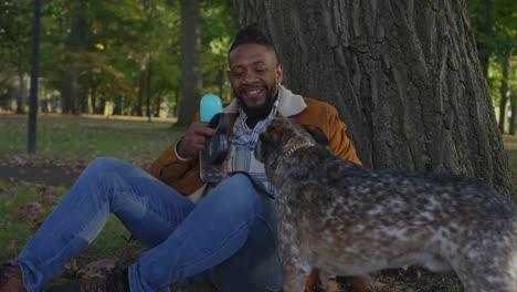 Un-Hombre-Negro-Tomando-Un-Descanso-Mientras-Pasea-A-Su-Perro-Mientras-Se-Sienta-Junto-A-Un-árbol-En-Un-Día-De-Otoño