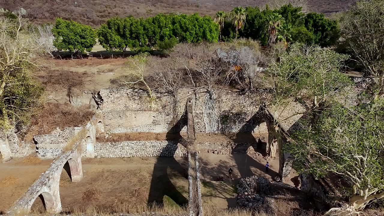 Dolly in drone flight over Ex Hacienda San Jacinto Ixtoluca ruins, La Mezquitera, Morelos. Overhead view of large stone ruins with arches and open walls surrounded by dry trees and greenery.
