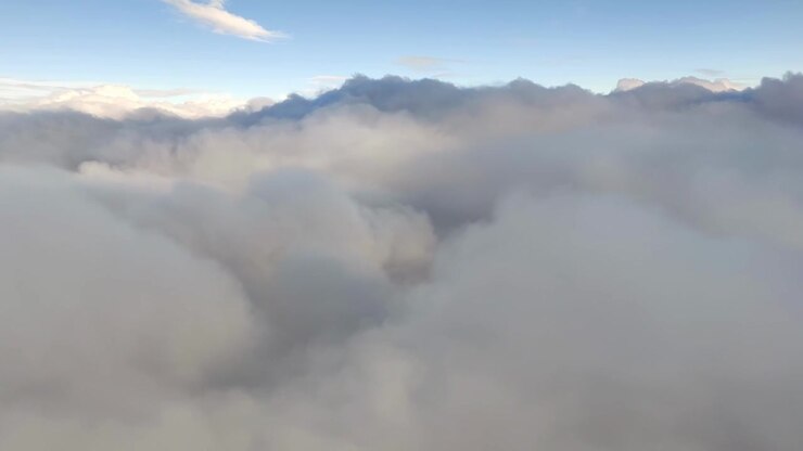 Plane moves close to huge fluffy clouds with sunlight and shadows. Blue sky visible above. Viewed from airplane window