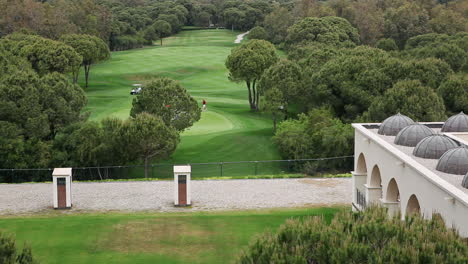 Breite-Aufnahme-Von-Golfspielern-Auf-Grünen-Feldern-Der-Golfplatzanlage-Mit-Caddy-Wagen-Und-Bäumen-Tagsüber