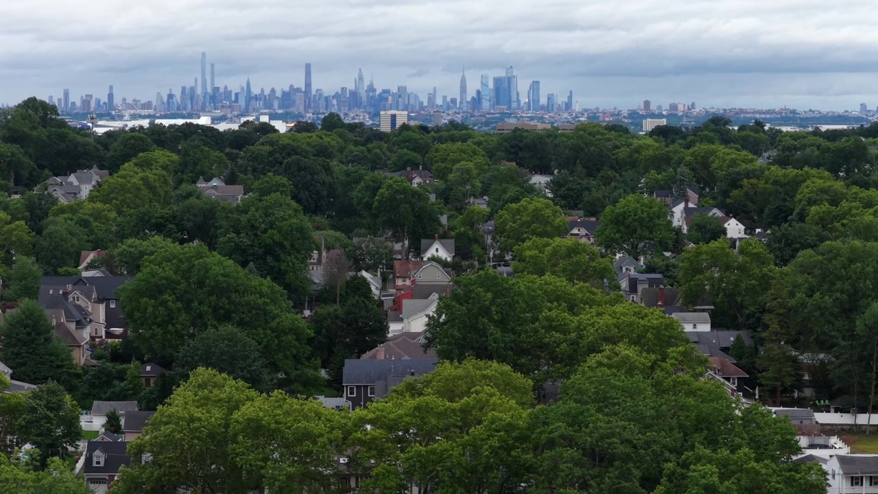 Tiheä vihreä puukatos peittää rauhallisia esikaupunkitaloja Cliftonissa, New Jerseyssä. Kaukainen sumuinen Manhattanin siluetti Empire State Buildingin kanssa kohoaa pilvisen, harmaan taivaan alla. Laaja ilmakuva dronella.