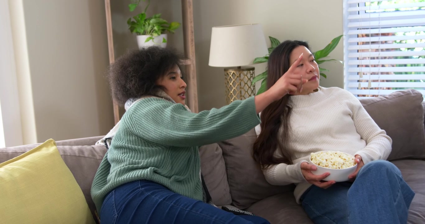 Afroamerikanske og asiatiske kvinder ser handling uden for skærmen på sofaen, mens de rækker ned i en popcornskål.