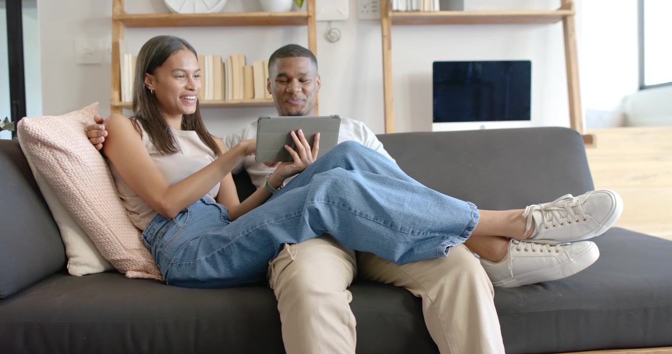 African American couple on sofa at home, man holding tablet while woman tapping showing content